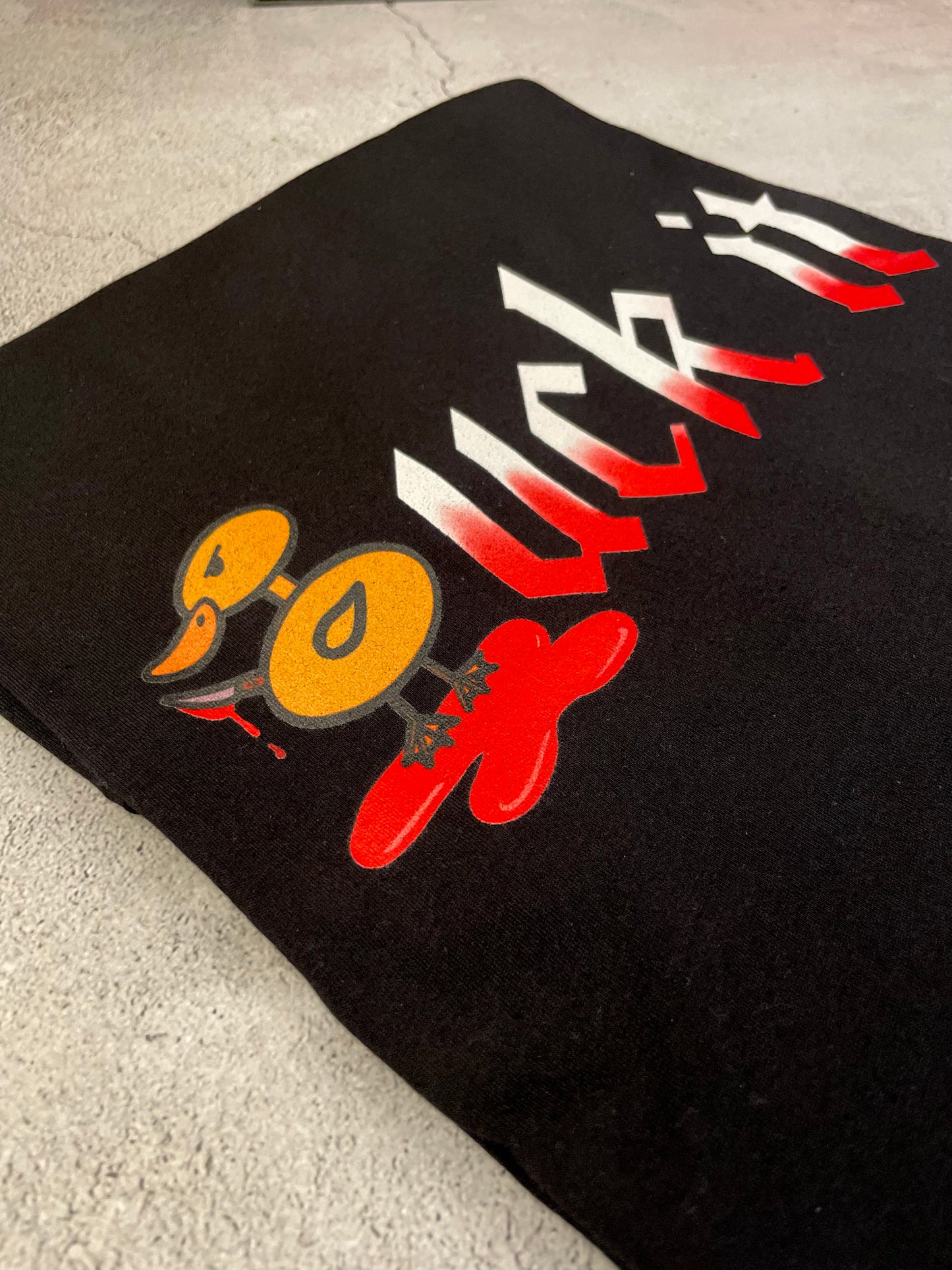 Angled close-up of a black t-shirt featuring the Duck It graphic design with a cartoon duck illustration and gothic-style lettering printed on the fabric.
