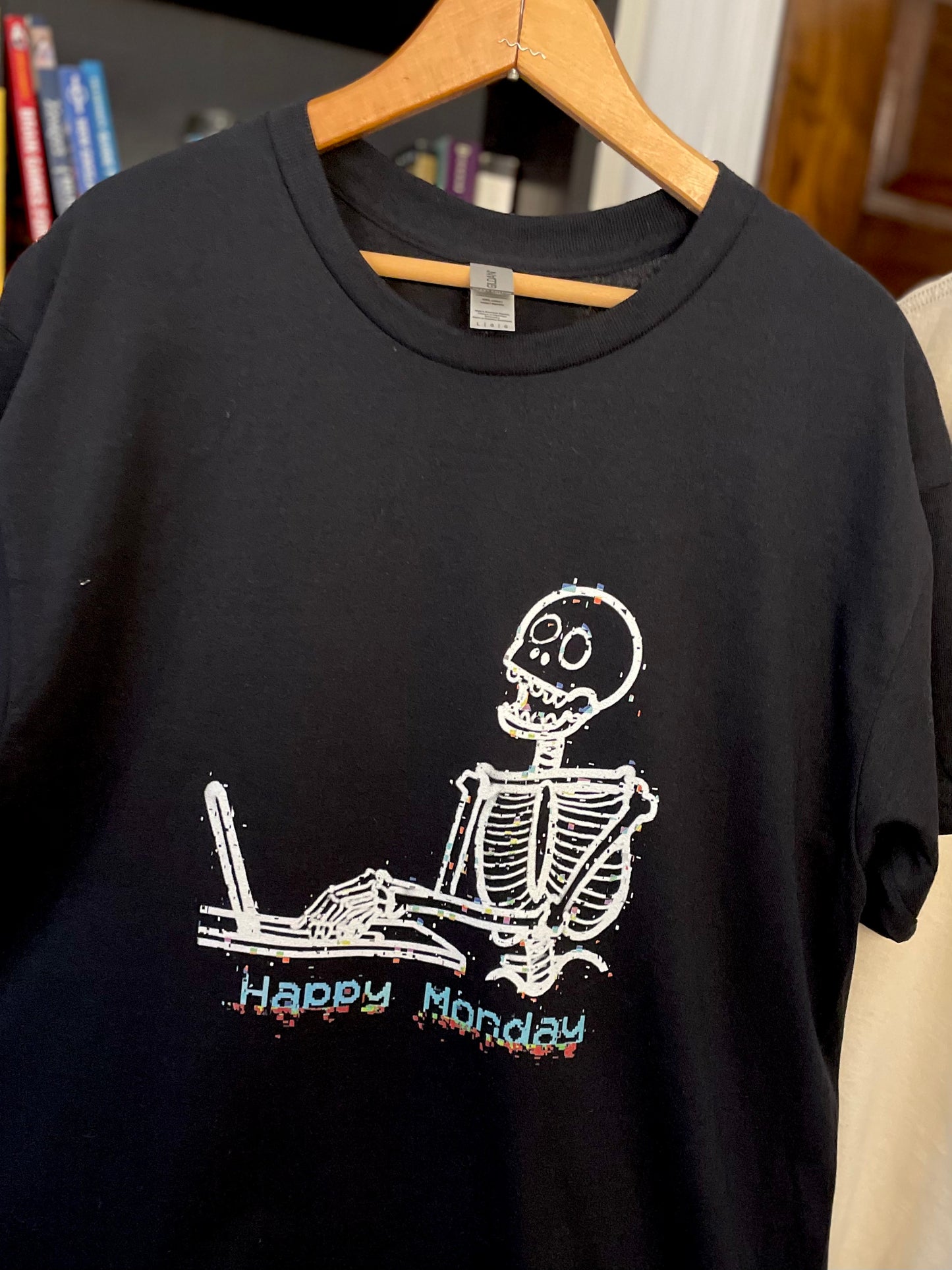 Real-life photo of a black t-shirt hanging on a hanger featuring a skeleton working on a laptop with the text “Happy Monday” underneath the illustration.