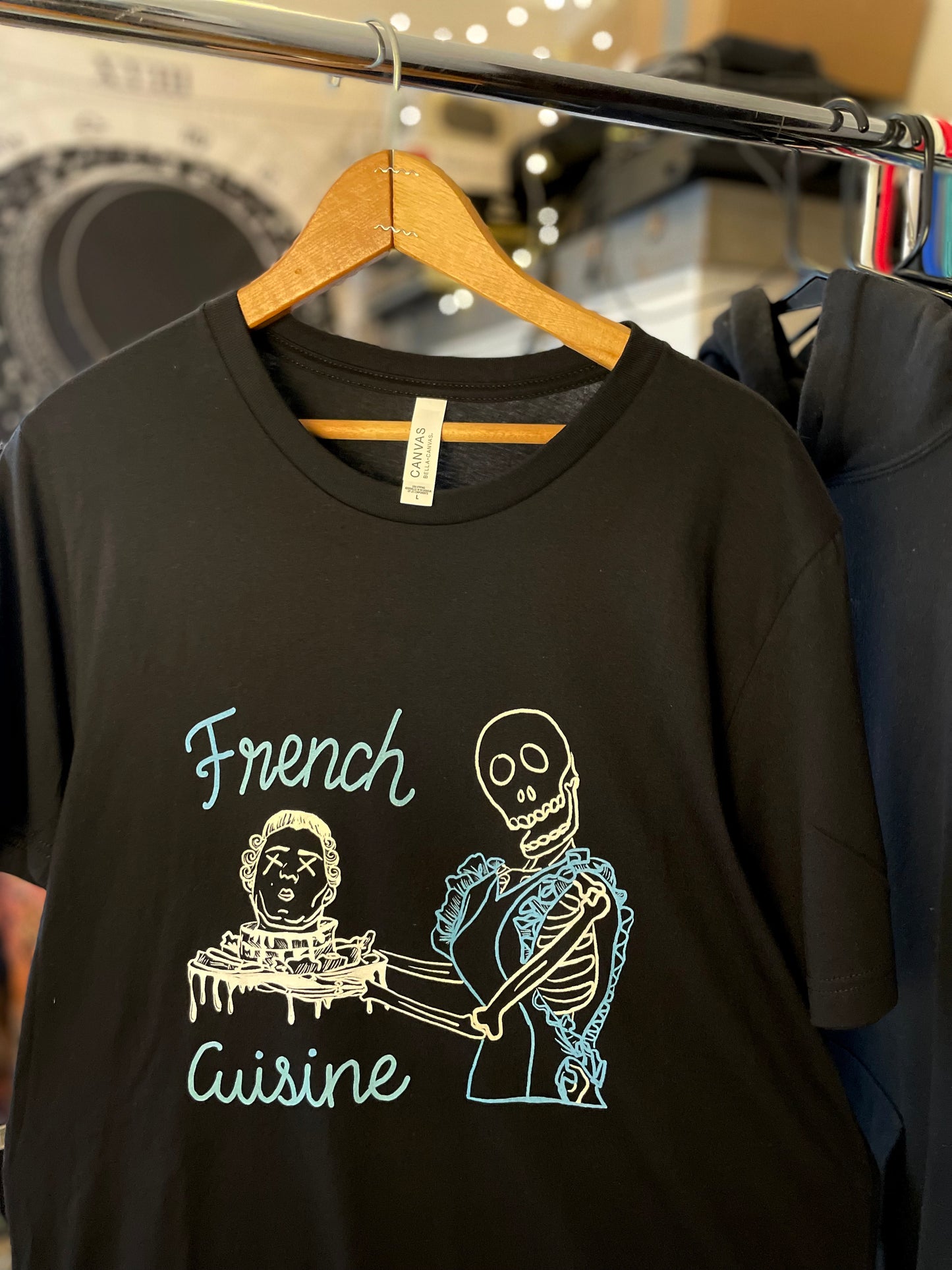 Black French Cuisine skeleton graphic tee hanging on rack in retail setting, dark humor shirt.