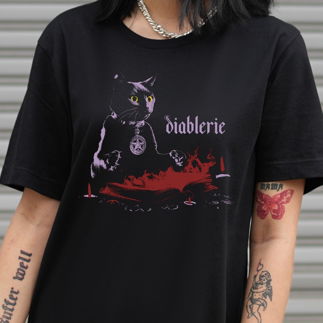 Model wearing the Dark Arts black cat spellbook t-shirt, shown from the chest up, with tattoos visible on the arms.